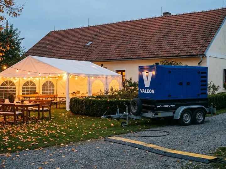 Zahrada venkovské usedlosti s bílým stanem a světelnými řetězy za soumraku, za plotem modrá elektrocentrála na přívěsu